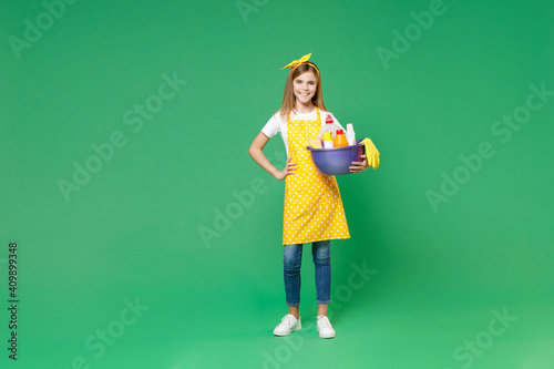 Full length funny little kid girl housewife 12-13 in apron stand with arm akimbo hold basin with detergent bottles washing cleansers doing housework isolated on green background. Housekeeping concept.