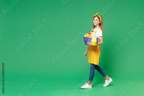 Full length side view smiling little kid girl housewife 12-13 in apron looking aside hold basin detergent bottles washing cleansers doing housework isolated on green background. Housekeeping concept. © ViDi Studio