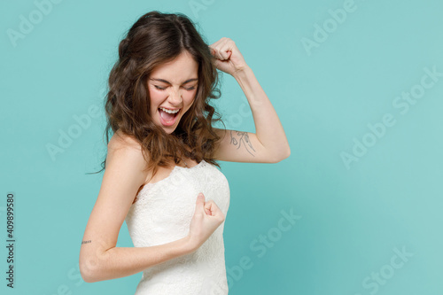 Side view of overjoyed bride young woman in white wedding dress doing winner gesture clenching fist say yes isolated on blue turquoise background studio portrait. Ceremony celebration party concept. © ViDi Studio