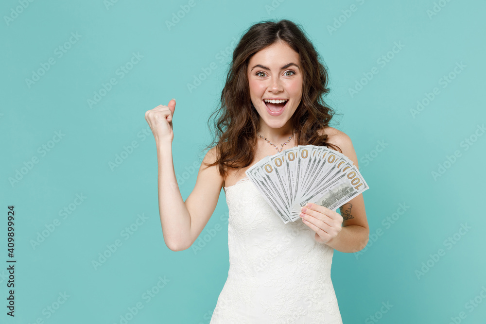 Happy bride young woman in white wedding dress hold fan cash money in dollar banknotes doing winner gesture isolated on blue turquoise background studio portrait. Ceremony celebration party concept. Happy bride young woman in white wedding dress hold fan cash money in dollar banknotes doing winner gesture isolated on blue turquoise background studio portrait. Ceremony celebration party concept.