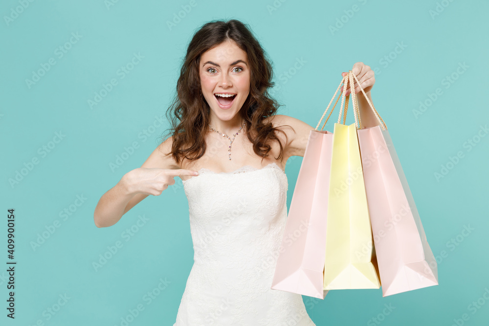 Excited bride young woman 20s in white wedding dress pointing index finger on package bags with purchases after shopping isolated on blue turquoise wall background. Ceremony celebration party concept. Excited bride young woman 20s in white wedding dress pointing index finger on package bags with purchases after shopping isolated on blue turquoise wall background. Ceremony celebration party concept.