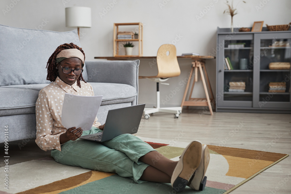 Full length portrait of modern African-American woman working from home while sitting on floor on graphic carpet and using laptop, copy space - obrazy, fototapety, plakaty 