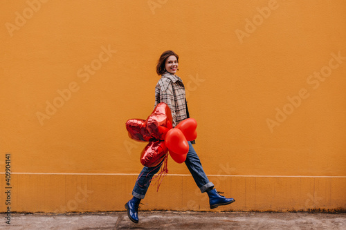 Full-length portrait of positive short-haired girl in jeans, boots Dr. Martins and tweed jacket. Woman with red balloons walks against tbackground of orange wall