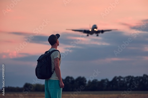 Young man looking up at flying airplane against moody sky during dusk. Themes nostalgia for travel and aviation. © Chalabala