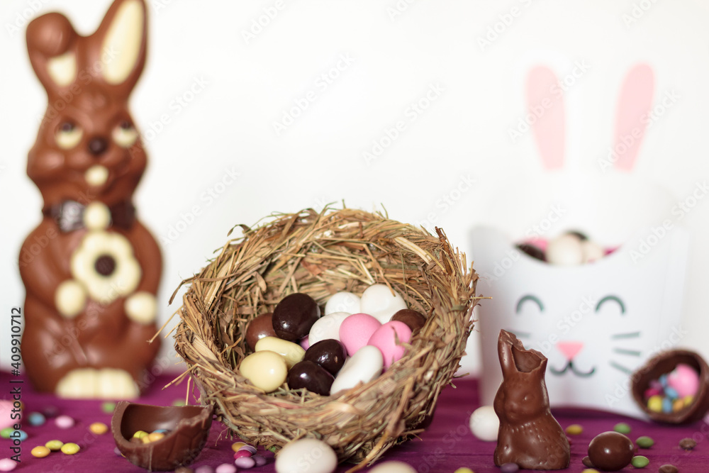 Bird nest with easter almonds, chocolate eggs, chocolate bunny and sweets on table against white background. Easter celebration concept