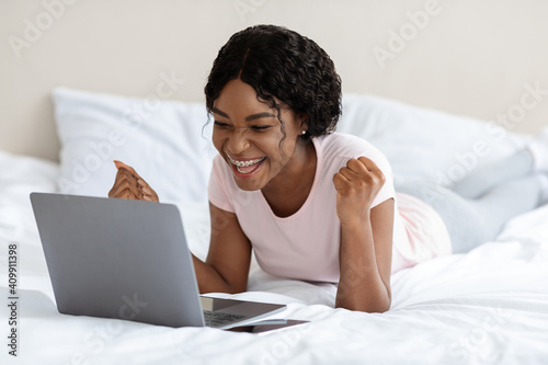 Positive african woman celebrating success, laying with laptop on bed