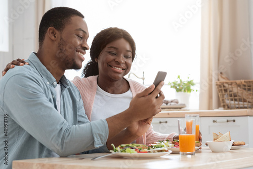 Cheerful black couple using smartphone while having breakfast in cozy kitchen