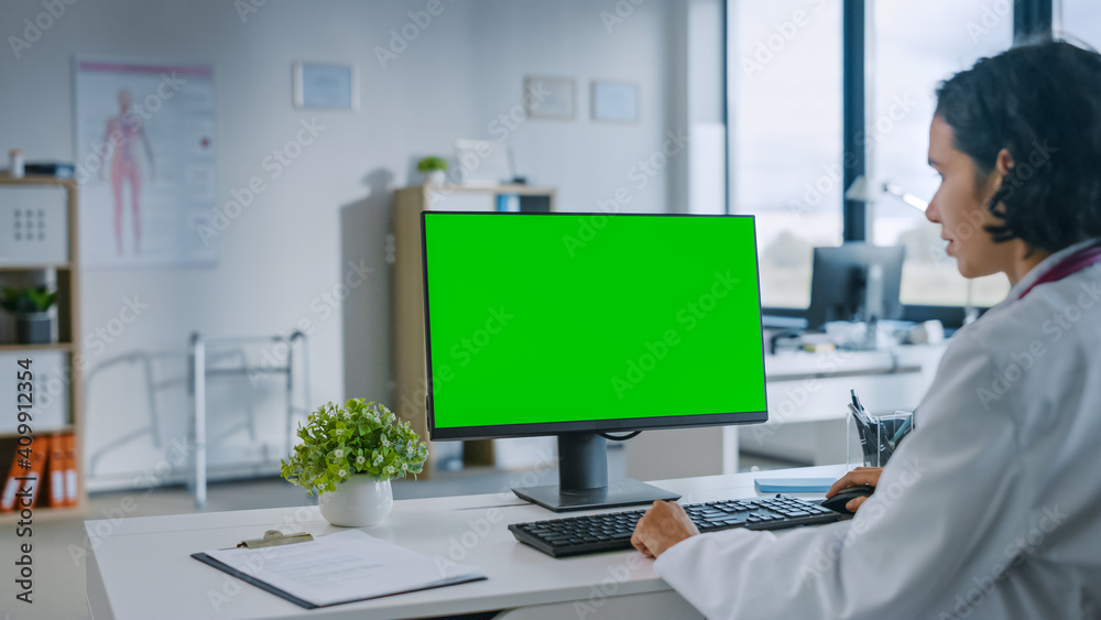 Female Medical Doctor is Working on a Computer with Green Screen Mock Up Display in a Health Clinic. Assistant in White Lab Coat is Reading Medical History Behind a Desk in Hospital Office. 