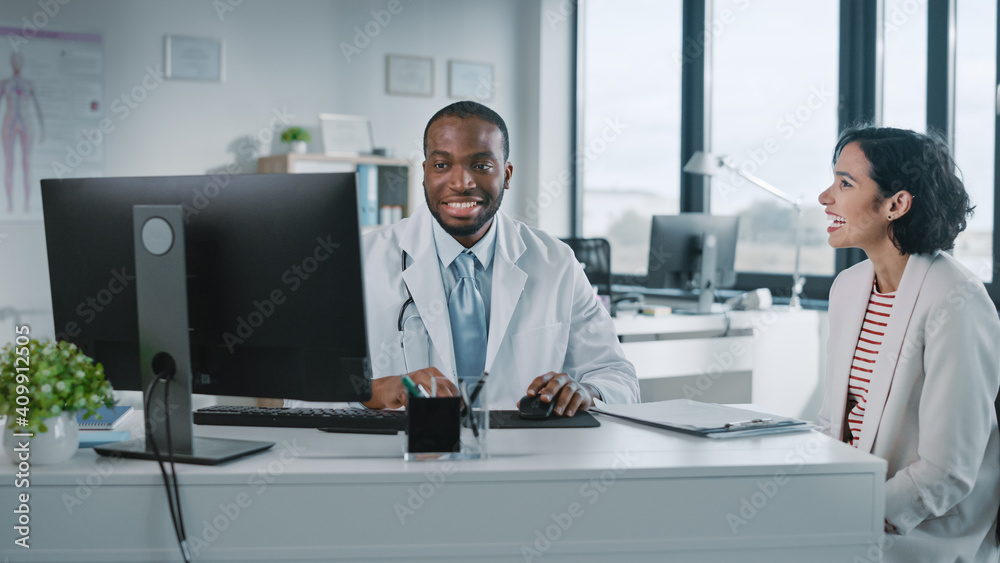 Black Medical Doctor is Reading Medical History of Female Patient and Speaking with Her During Consultation in a Health Clinic. Physician in Lab Coat is Sitting Behind a Computer in Hospital Office.