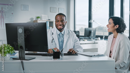 Black Medical Doctor is Reading Medical History of Female Patient and Speaking with Her During Consultation in a Health Clinic. Physician in Lab Coat is Sitting Behind a Computer in Hospital Office.