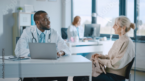 Family Doctor is Reading Medical History of Senior Female Patient and Speaking with Her During Consultation in a Health Clinic. Physician in Lab Coat Sitting Behind a Laptop in Hospital Office. 