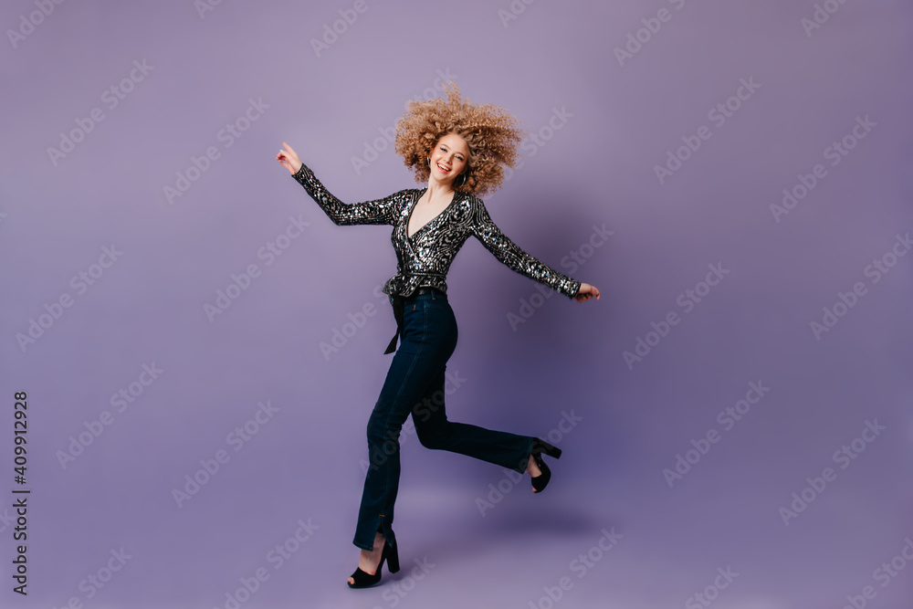 Positive blonde woman happily jumping on purple background. Girl dressed in silver blouse and jeans laughs