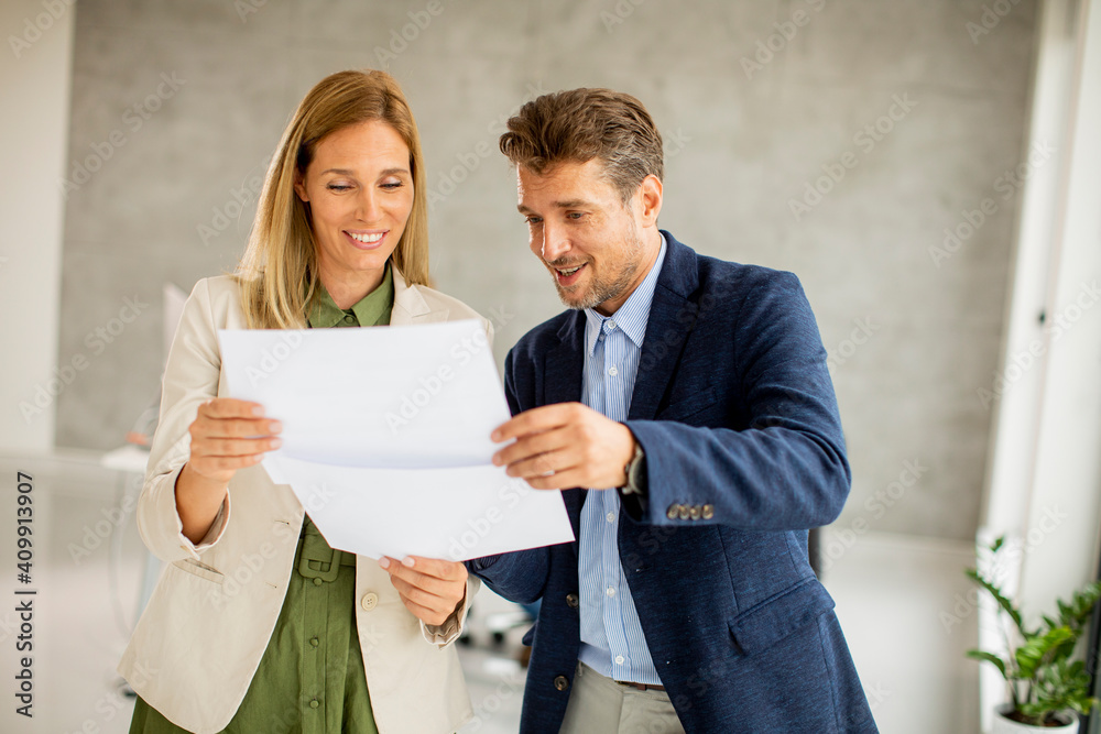 Man and woman discussing with paper in hands indoors in the office with young people works behind them Man and woman discussing with paper in hands indoors in the office with young people works behind them