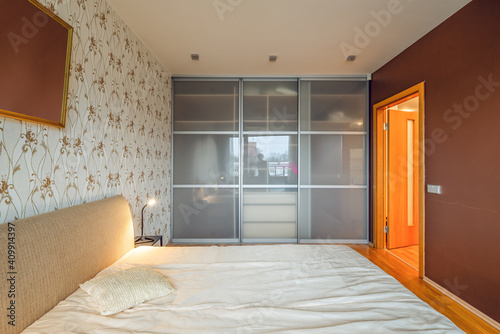 Contemporary interior of bedroom in luxury flat. Side view of cozy bed. Huge sliding door wardrobe. Lamp on nightstand.