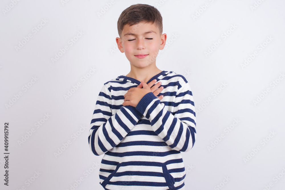 little cute boy kid wearing stripped t-shirt closes eyes and keeps hands on chest near heart, expresses sincere emotions, being kind hearted and honest. Body language and real feelings concept. little cute boy kid wearing stripped t-shirt closes eyes and keeps hands on chest near heart, expresses sincere emotions, being kind hearted and honest. Body language and real feelings concept.