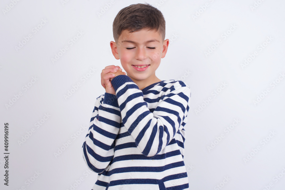 Dreamy little cute boy kid wearing stripped t-shirt against white wall with pleasant expression, closes eyes, keeps hands crossed near face, thinks about something pleasant Dreamy little cute boy kid wearing stripped t-shirt against white wall with pleasant expression, closes eyes, keeps hands crossed near face, thinks about something pleasant