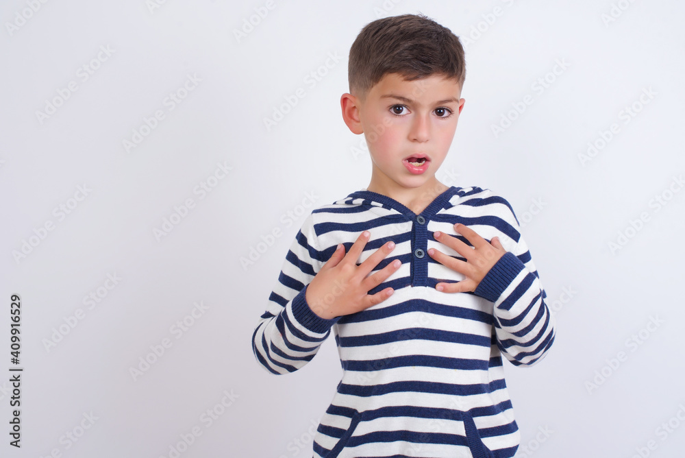 little cute boy kid wearing stripped t-shirt against white wall keeps hands on chest feeling shocked and scared, mouth widely opened, stares at camera saying: Who, me? little cute boy kid wearing stripped t-shirt against white wall keeps hands on chest feeling shocked and scared, mouth widely opened, stares at camera saying: Who, me?