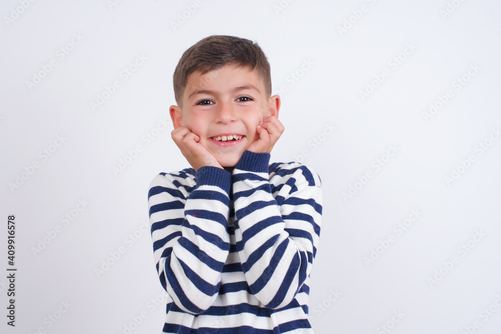 Dreamy little cute boy kid wearing red stripped t-shirt against white wall keeps hands pressed together under chin, looks with happy expression, has toothy smile. Dreamy little cute boy kid wearing red stripped t-shirt against white wall keeps hands pressed together under chin, looks with happy expression, has toothy smile.