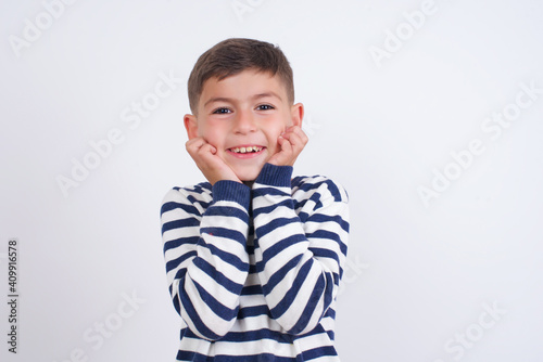 Dreamy little cute boy kid wearing red stripped t-shirt against white wall keeps hands pressed together under chin, looks with happy expression, has toothy smile.