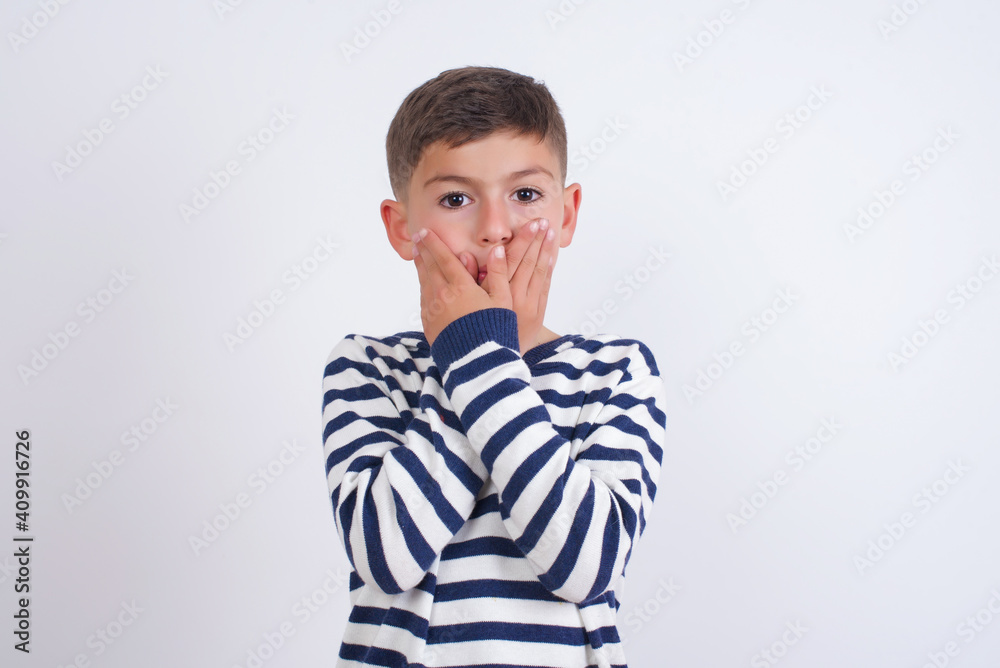 Upset little cute boy kid wearing red stripped t-shirt against white wall, covering her mouth with both palms to prevent screaming sound, after seeing or hearing something bad. Upset little cute boy kid wearing red stripped t-shirt against white wall, covering her mouth with both palms to prevent screaming sound, after seeing or hearing something bad.