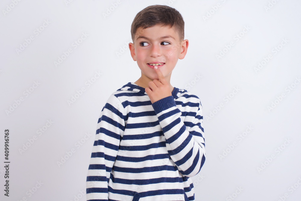 little cute boy kid wearing red stripped t-shirt against white wall with thoughtful expression, looks to the camera, keeps hand near face, bitting a finger thinks about something pleasant. little cute boy kid wearing red stripped t-shirt against white wall with thoughtful expression, looks to the camera, keeps hand near face, bitting a finger thinks about something pleasant.