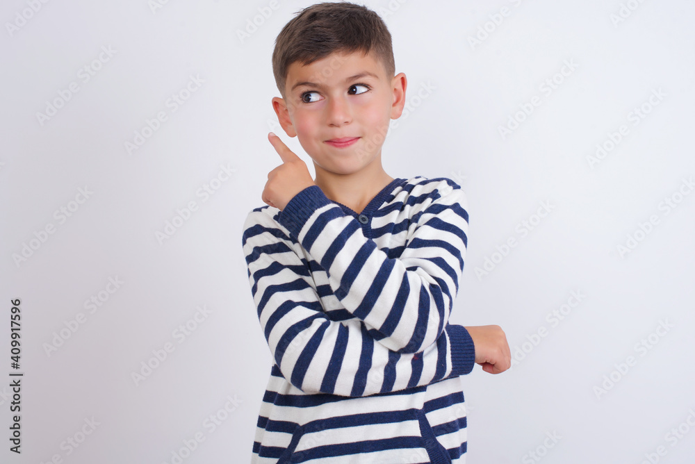 Portrait of little cute boy kid wearing red stripped t-shirt against white wall posing on camera with tricky look, presenting product with index finger. Advertisement concept. Portrait of little cute boy kid wearing red stripped t-shirt against white wall posing on camera with tricky look, presenting product with index finger. Advertisement concept.