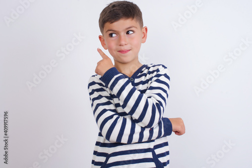 Portrait of little cute boy kid wearing red stripped t-shirt against white wall posing on camera with tricky look, presenting product with index finger. Advertisement concept.