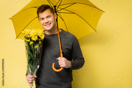 Smiling man with flowers and umbrella looking at camera on yellow background