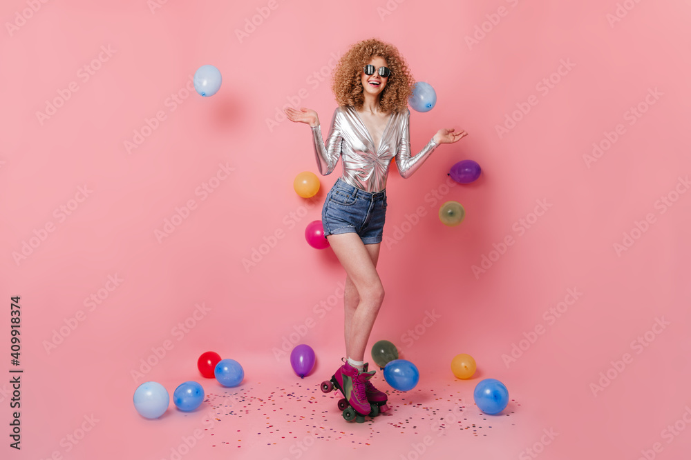 Fototapeta premium Cheerful curly woman riding roller skates. Lady in sunglasses dressed in silver top and denim shorts posing on pink background with balloons
