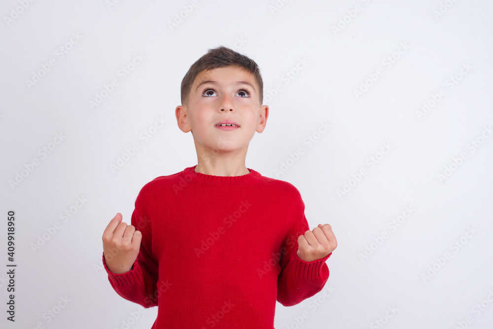 Little cute boy kid wearing red knitted sweater against white wall having success being glad to achieve his goals. Victory and triumph concept. Little cute boy kid wearing red knitted sweater against white wall having success being glad to achieve his goals. Victory and triumph concept.