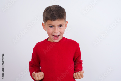 Little cute boy kid wearing red knitted sweater against white wall raising fists up screaming with joy being happy to achieve goals.