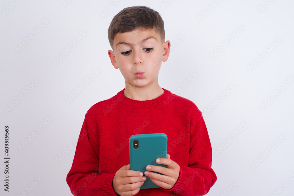 Little cute boy kid wearing red knitted sweater against white wall looks with bugged eyes, holds modern smart phone, receives unexpected message from friend, reads reminder. Little cute boy kid wearing red knitted sweater against white wall looks with bugged eyes, holds modern smart phone, receives unexpected message from friend, reads reminder.