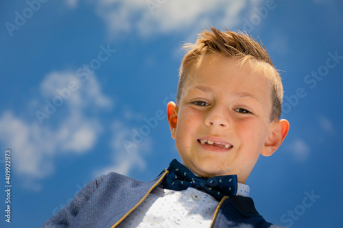 Tablou pe pânză Image of a cute smiling young boy looking down at the camera with the blue sky spotted with white clouds as a background