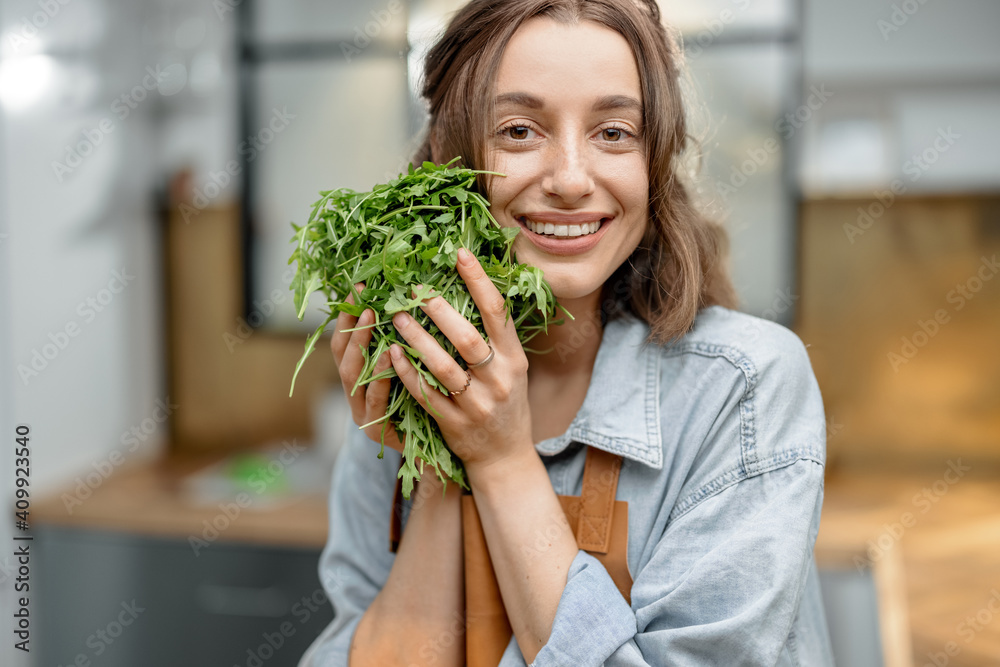 Portrait of pretty smiling woman in apron with fresh arugula on the kitchen. Healthy cooking concept. Close up. High quality photo - obrazy, fototapety, plakaty 