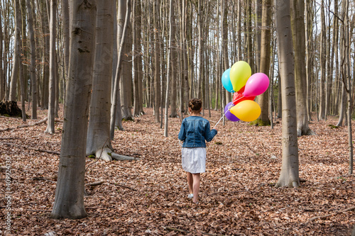 Young girl, seen from behind, walking in the woods with a bunch of balloons.