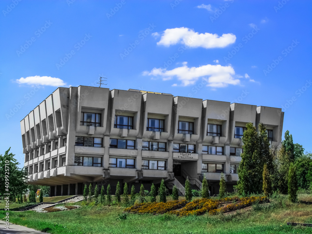 Sumy Regional Universal Scientific Library building on a sunny summer day (Ukraine). Gray concrete modern building with large windows amidst green park with flowerbed