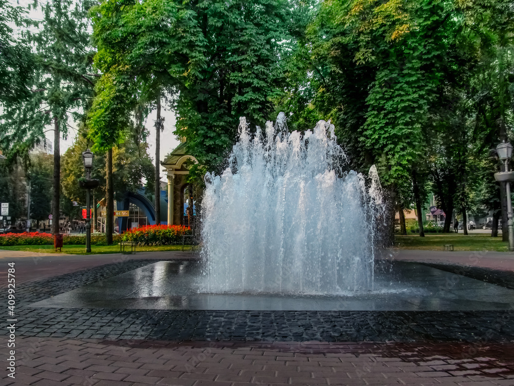 Fountain Sugar in Pokrovsky Square in Sumy (Ukraine). Modern fountain with jets of water gushing from the floor against the backdrop of greenery and flowers in the park