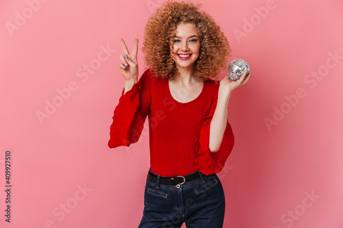 Excited girl dressed in red blouse and jeans posing with disco ball and showing sign of peace on pink background