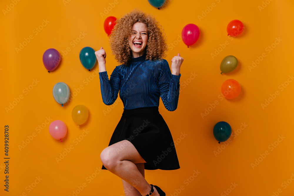 Fototapeta premium Active girl in black skirt and blue blouse makes winning gesture and laughs, posing on yellow background with balloons