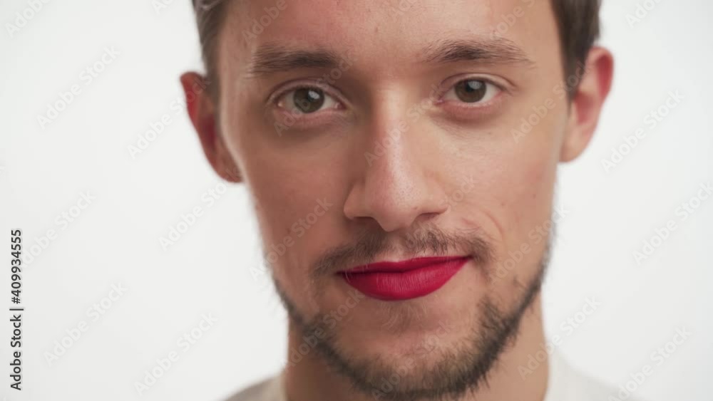 Young attractive American boy wear make-up – red lipstick on lips, with beard, moustache smile, flirt, raises his eyebrows up isolated on white background close-up in slow motion. Fashion model portra Young attractive American boy wear make-up – red lipstick on lips, with beard, moustache smile, flirt, raises his eyebrows up isolated on white background close-up in slow motion. Fashion model portra
