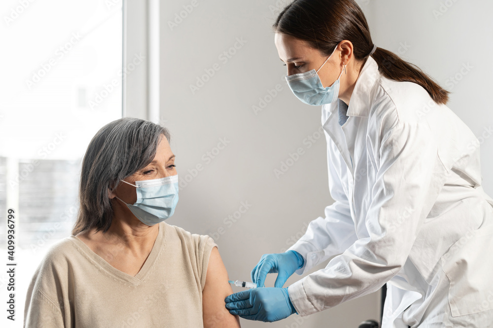 A nurse, doctor in medical gown giving injection to senior mature woman at hospital, at home. Medic giving injection of Covid-19 or coronavirus vaccine to a female senior patient A nurse, doctor in medical gown giving injection to senior mature woman at hospital, at home. Medic giving injection of Covid-19 or coronavirus vaccine to a female senior patient