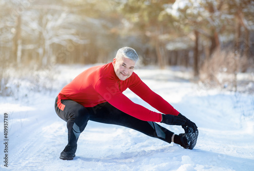 Outdoor winter activities concept. Joyful senior man stretching his legs before jogging at snowy park on cold day