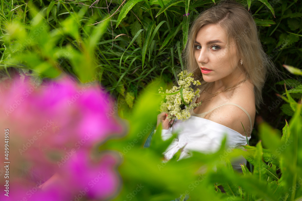 Sensual blonde girl in a white shirt stands in high grass with purple flowers and holds flowers in her hand on a summer day. High quality photo