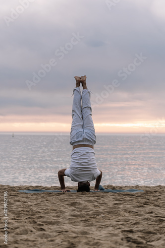 Healthy man in white clothes practising yoga exercises in the beach. Person doing Vinyasa yoga stretching series