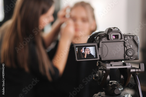 Woman make makeup for attractive blond hair woman in studio. Selective focus of digital camera with makeup artist applying cosmetics for beautiful model © Aleksandr