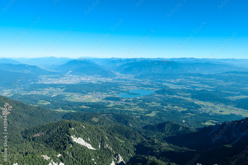 A panoramic view on the Alps from the top of Mittagskogel in Austrian Alps. Clear and sunny day. There is a lake at the bottom. A bit of haze in the valley. Outdoor activity. Alpine mountain chains