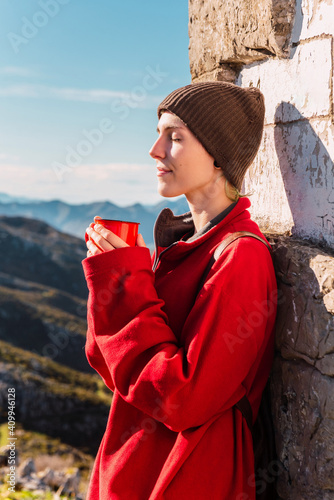 Side view of androgynous traveler standing in highlands on sunny day and enjoying hot drink during trip in El Mazuco with eyes closed photo