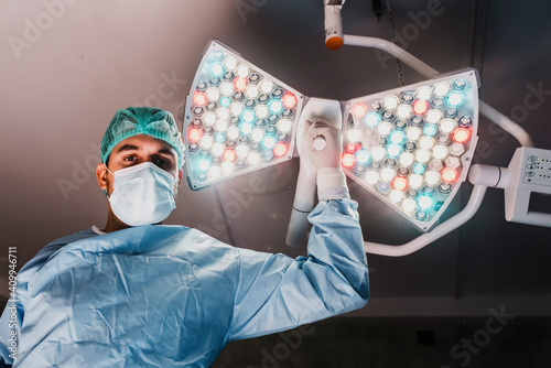 From below of serious male surgeon in protective mask and medical uniform adjusting surgical lamp while preparing for operation in modern operating theater photo