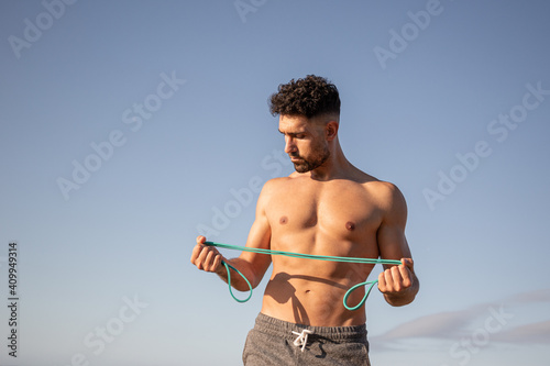 Muscular male bodybuilder with naked torso standing on seashore and doing exercises with resistance band during workout in summer