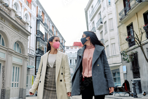 Two happy women talking and walking in the street with their face masks on. © David